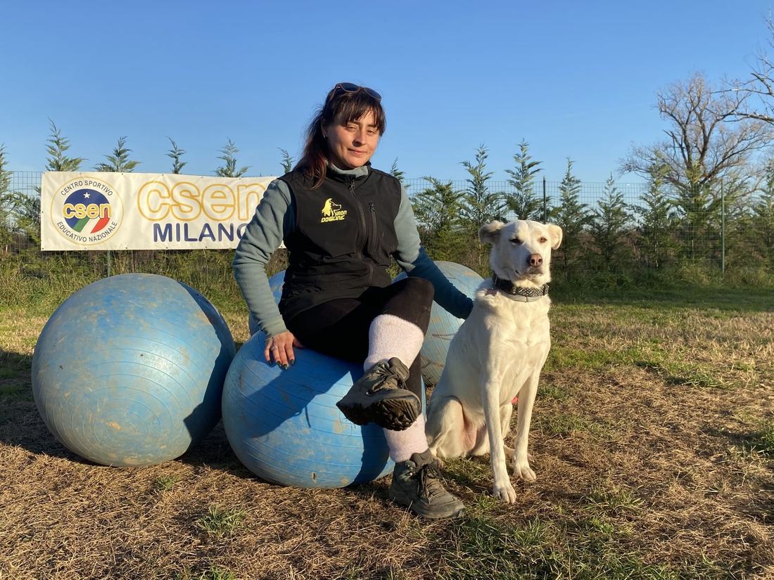 Un'istruttrice seduta su un grande pallone azzurro accanto a un cane bianco, con lo striscione CSEN Milano